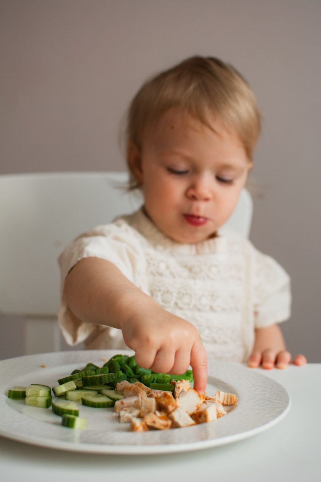 A little girl picking food from a plate of cucumber, greens and chicken