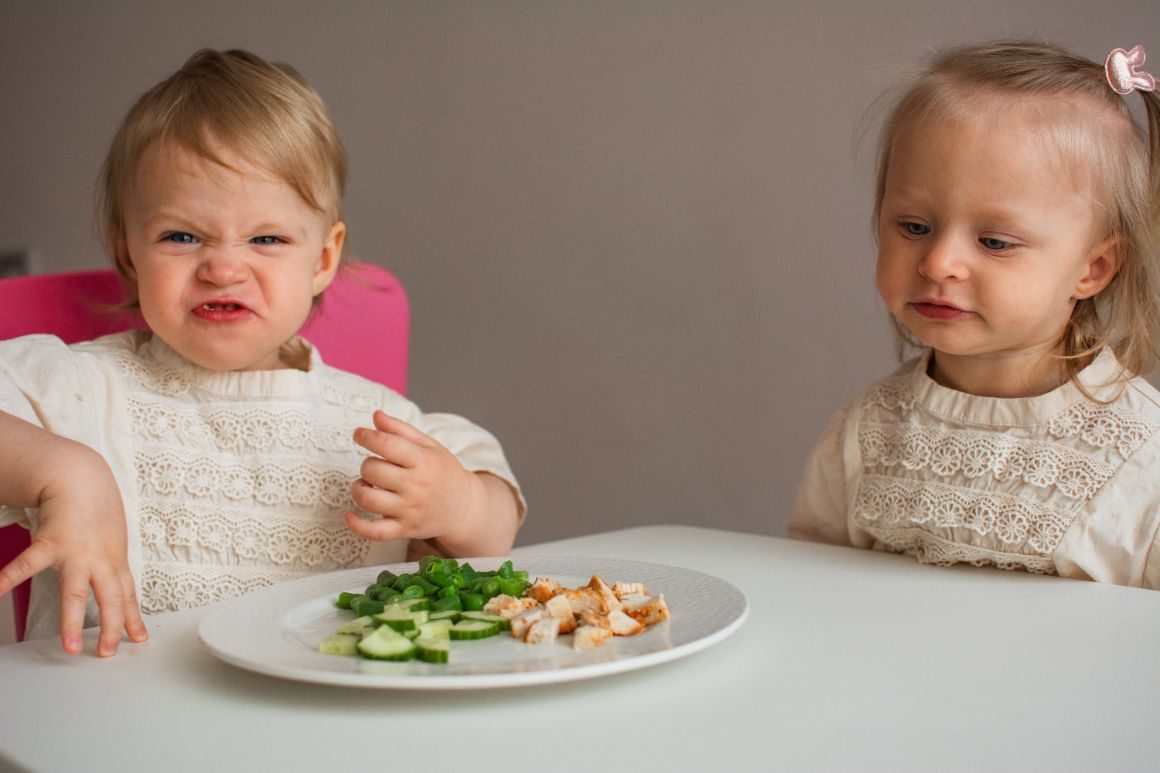 A little girl grimacing and refusing a plate of cucumber, greens and chicken