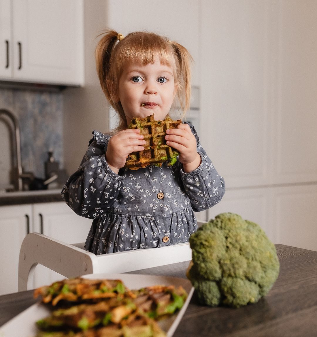 A little girl in a kitchen eating vegetable waffles, with broccoli beside her