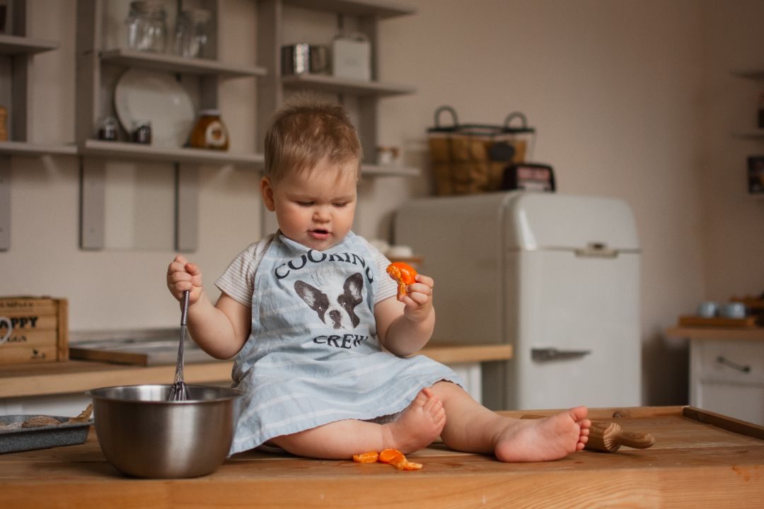 A baby sitting on a kitchen counter with a whisk and a piece of carrot