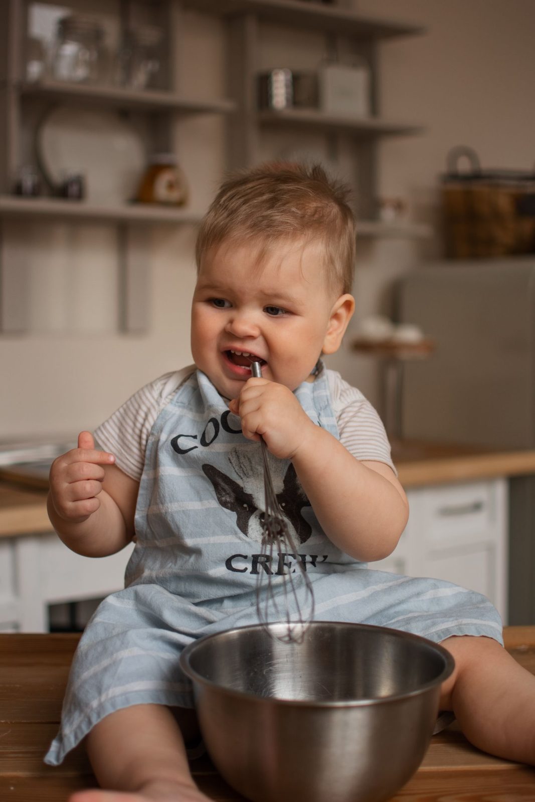 A smiling baby in an apron sitting in the kitchen with a bowl and a whisk